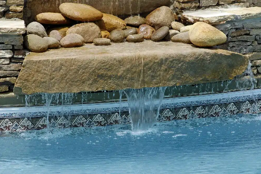 A serene poolside waterfall cascades over a smooth stone ledge into a bright blue pool. The scene conveys tranquility and relaxation.